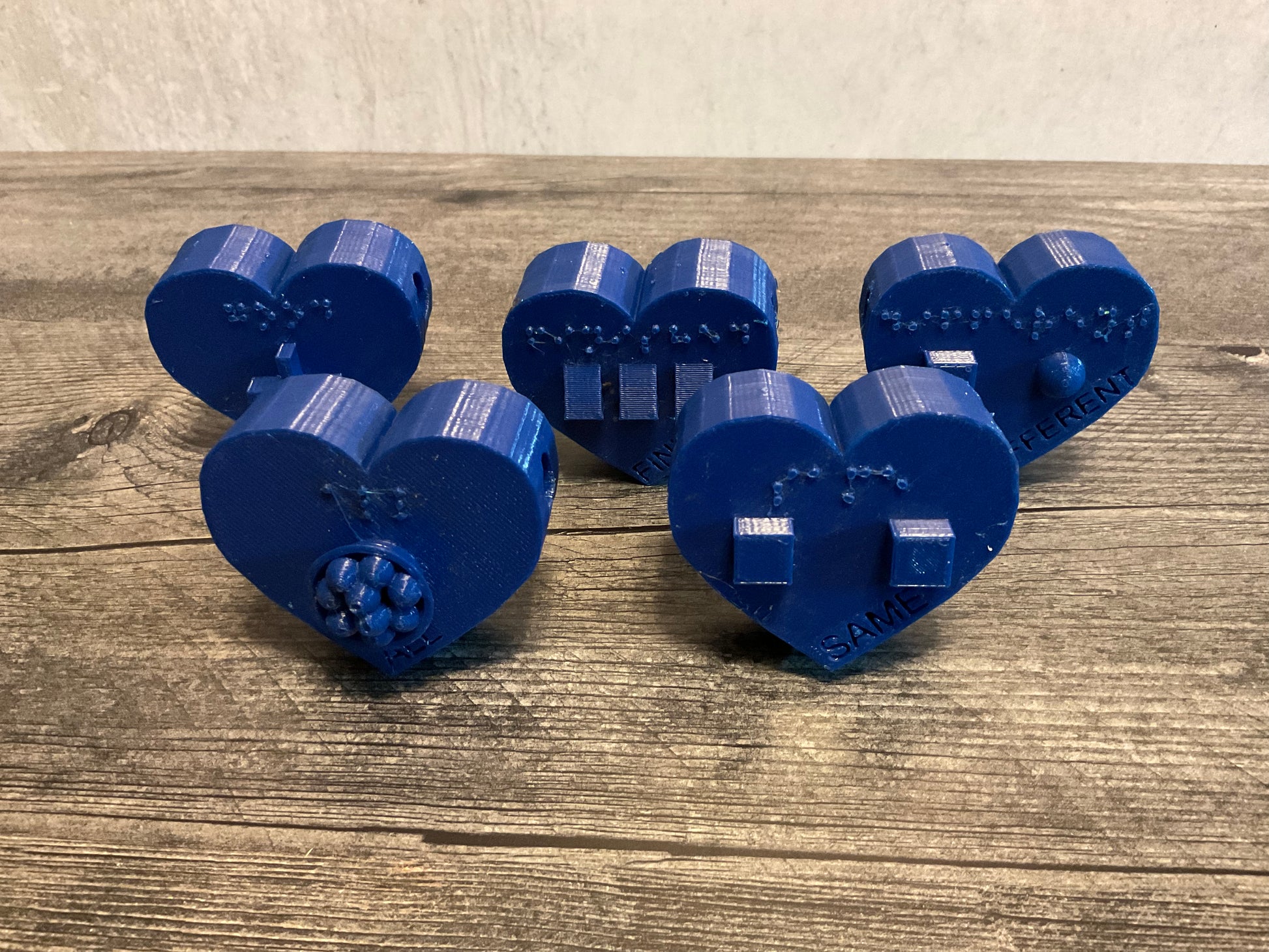 five blue heart shaped beads. Each has it's own symbol and braille as well as print Good Same Finished All Different