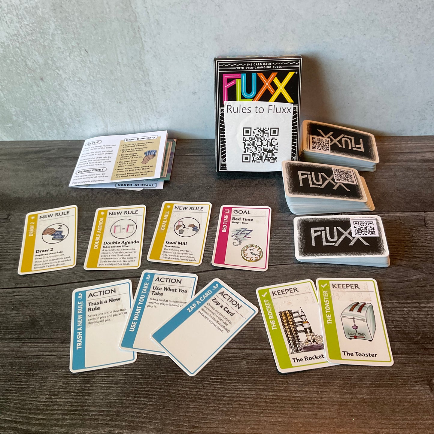 Wide shot of the game. The QR codes on the back of the cards can be seen. A few action, rules, and keepers are shown. Most of the function of the cards is embossed with transparent braille but the QR codes provide the original text.