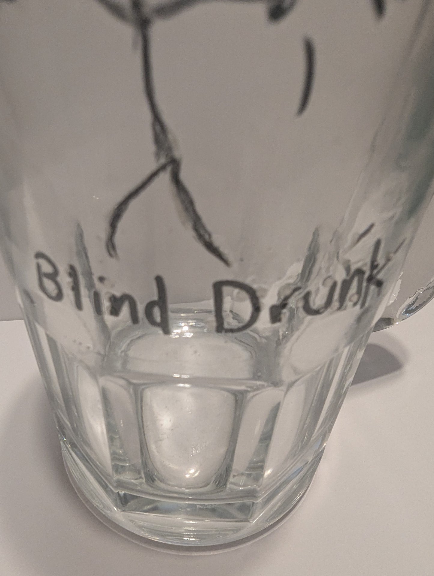 A close-up of a clear glass mug with a black hand-drawn tactile stick figure and the words 'Blind Drunk' written on it in both print and braille.