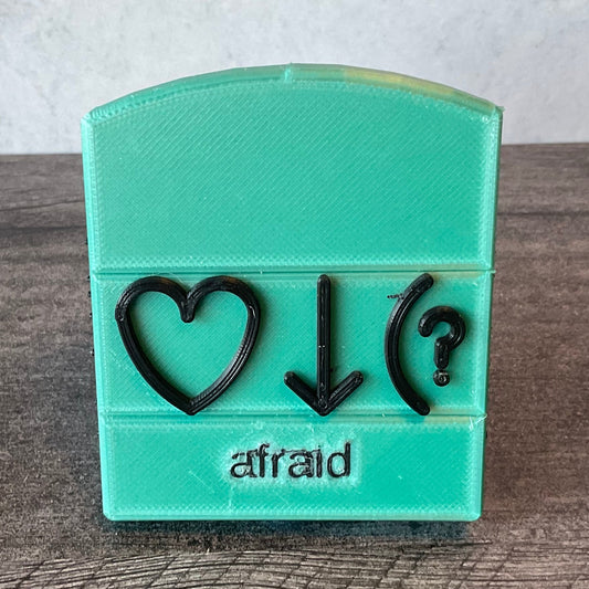 Afraid bliss symbol. This is an example but all words are this same design. Front view. Base is color is teal. Front says afraid in print and has the 3 symbols that Volk uses for this word, heart, down arrow and parenthesis with question mark.