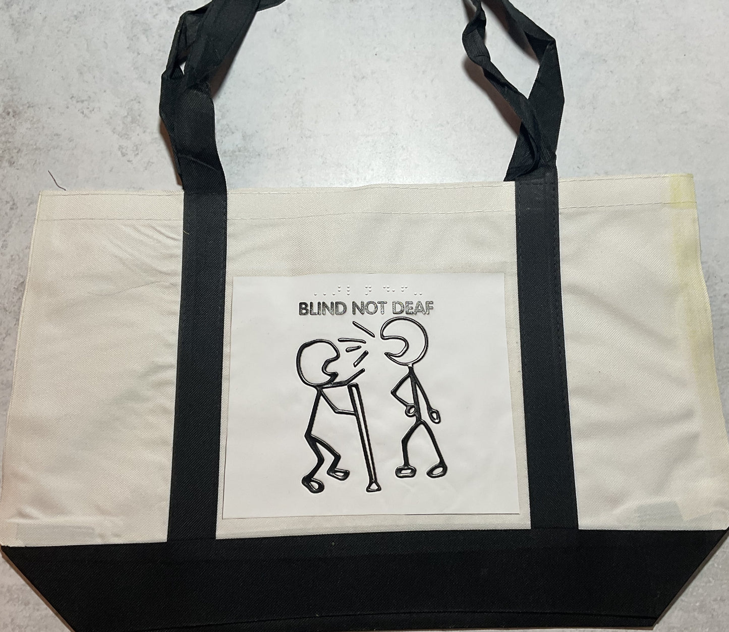 Wide shot of tote bag with a stick figure screaming at a blind person with the phrase "Blind not deaf" written in both print and braille.