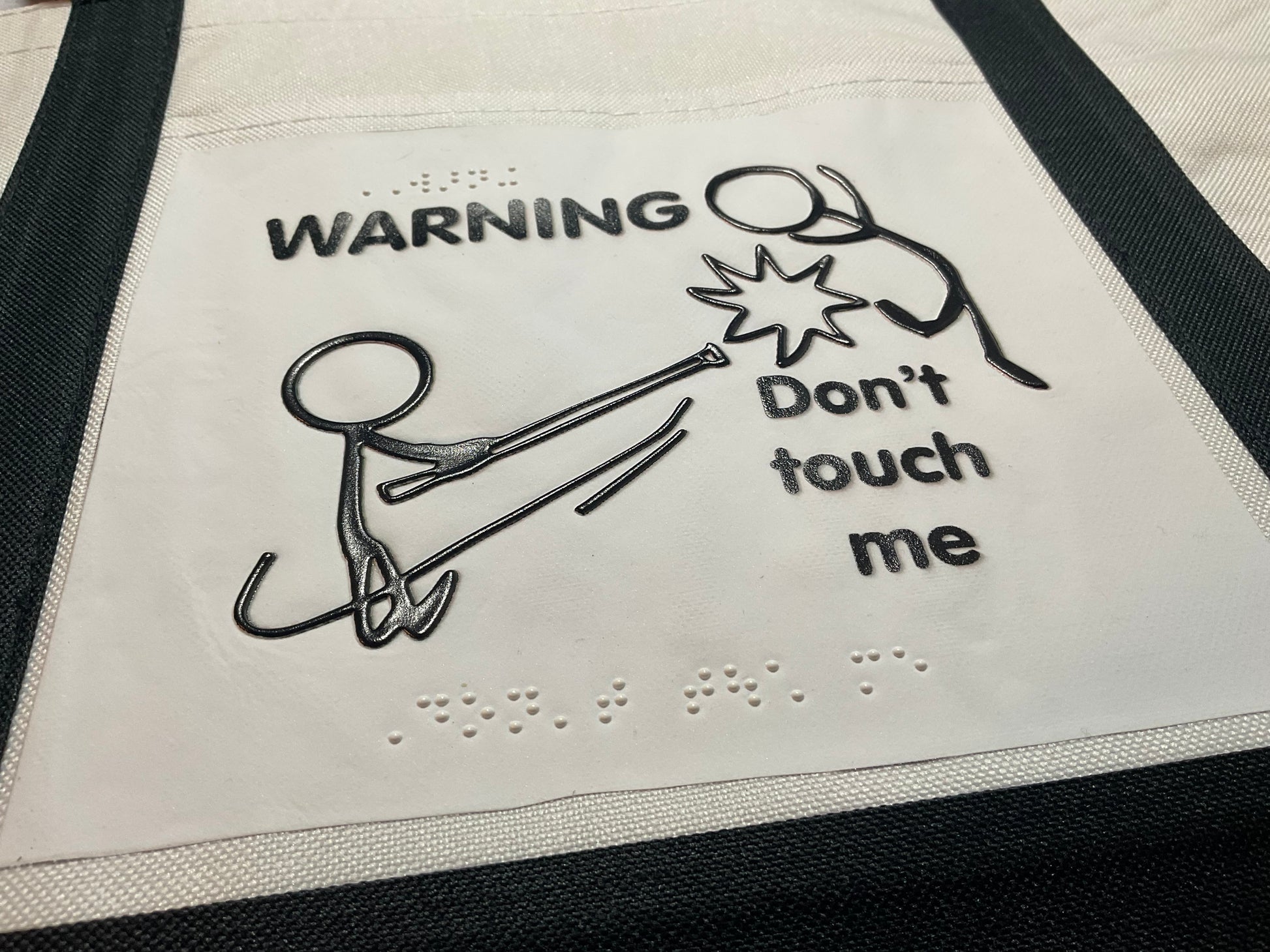 Close up on tote bag showing the texture and the braille on the tote bag. It says Warning, don't touch me in both print and braille.