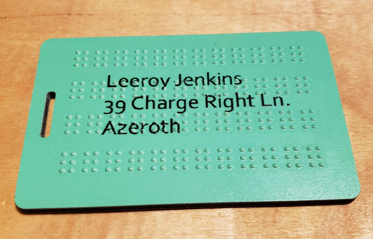 A picture of a green luggage tag with 4 lines of braille on it. Print lettering shows the address on top of the braille.