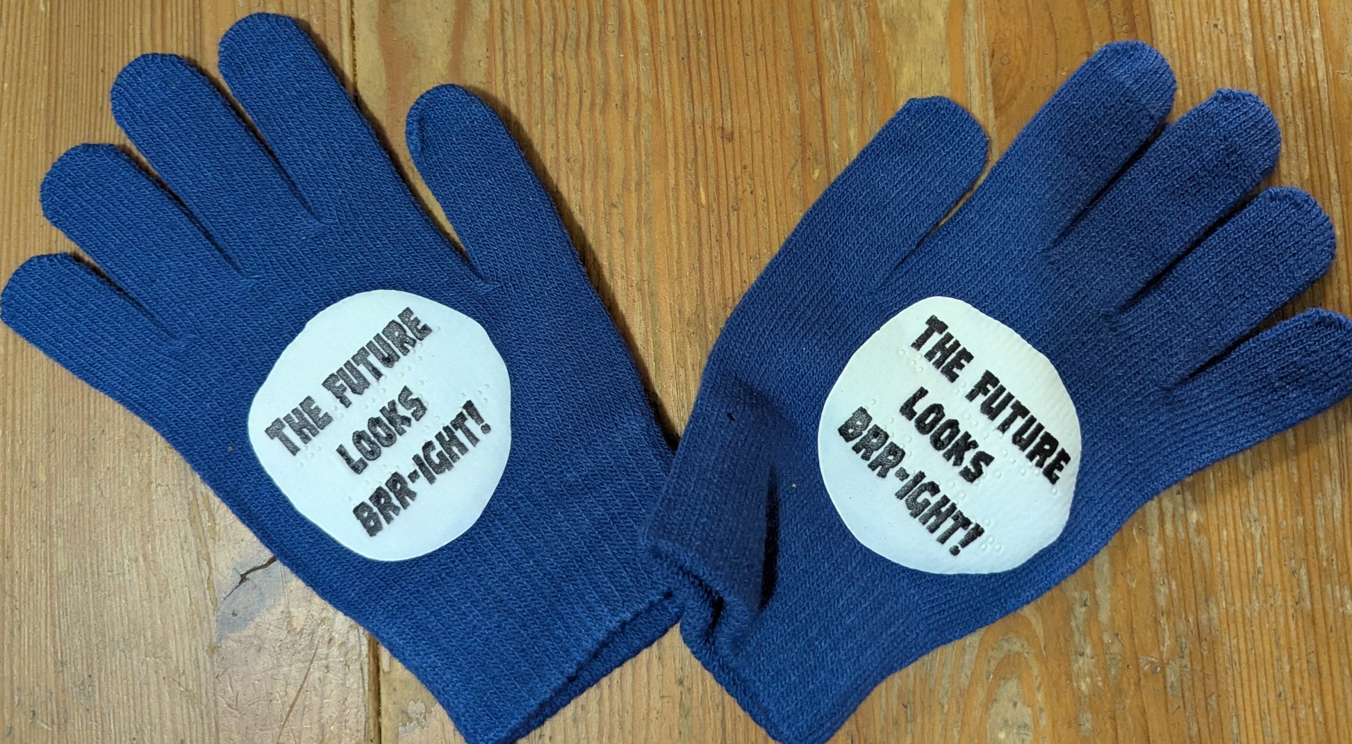 Pair of blue gloves with the print of a snowball on them with the words "The Future Looks Brr-ight!" in both print and braille on a wooden surface