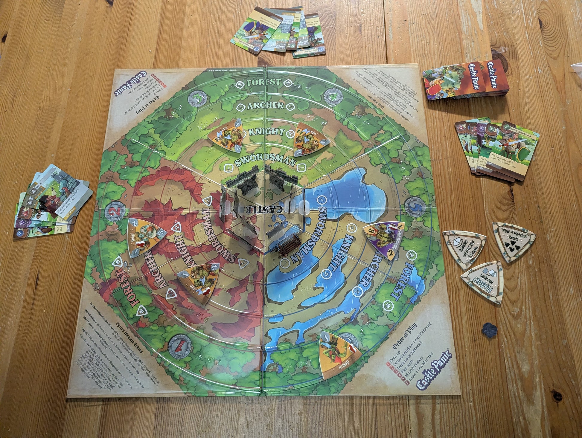 Board game setup on a wooden table with pieces and cards. The board has a clear thermoform on it with braille on it and the monster tokens are all replaced with 150% scale tokens with braille on them.