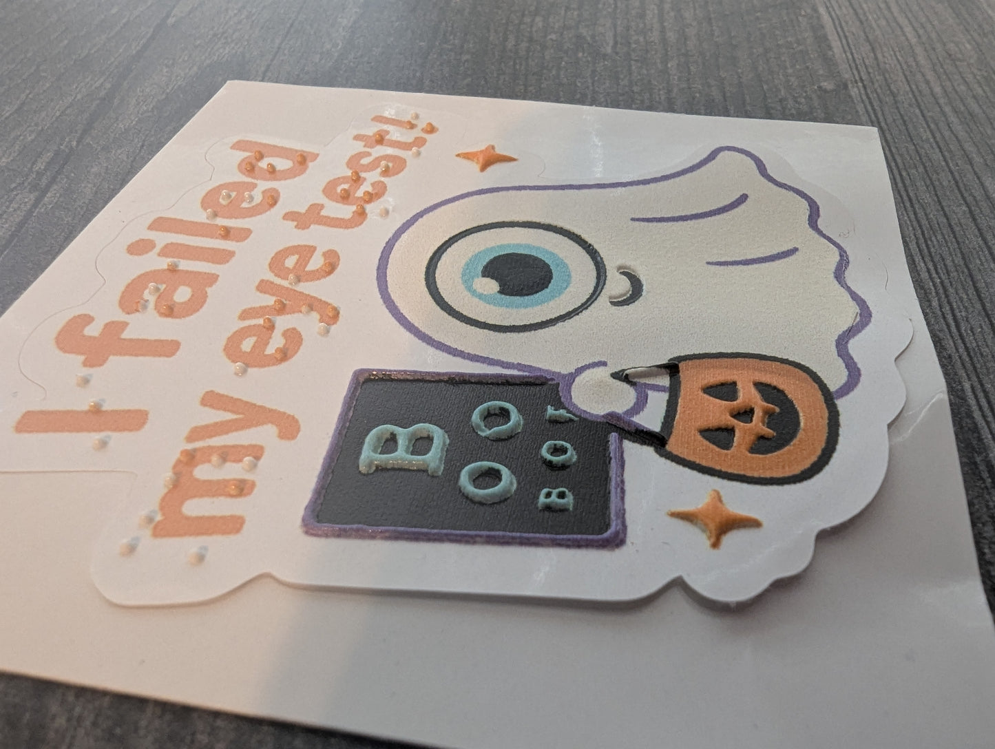 Kiss cut sticker with cartoon eye and text 'I failed my eye test!' on a gray surface with both braille and print. The eye, ghost, eye chart and more are tactile.