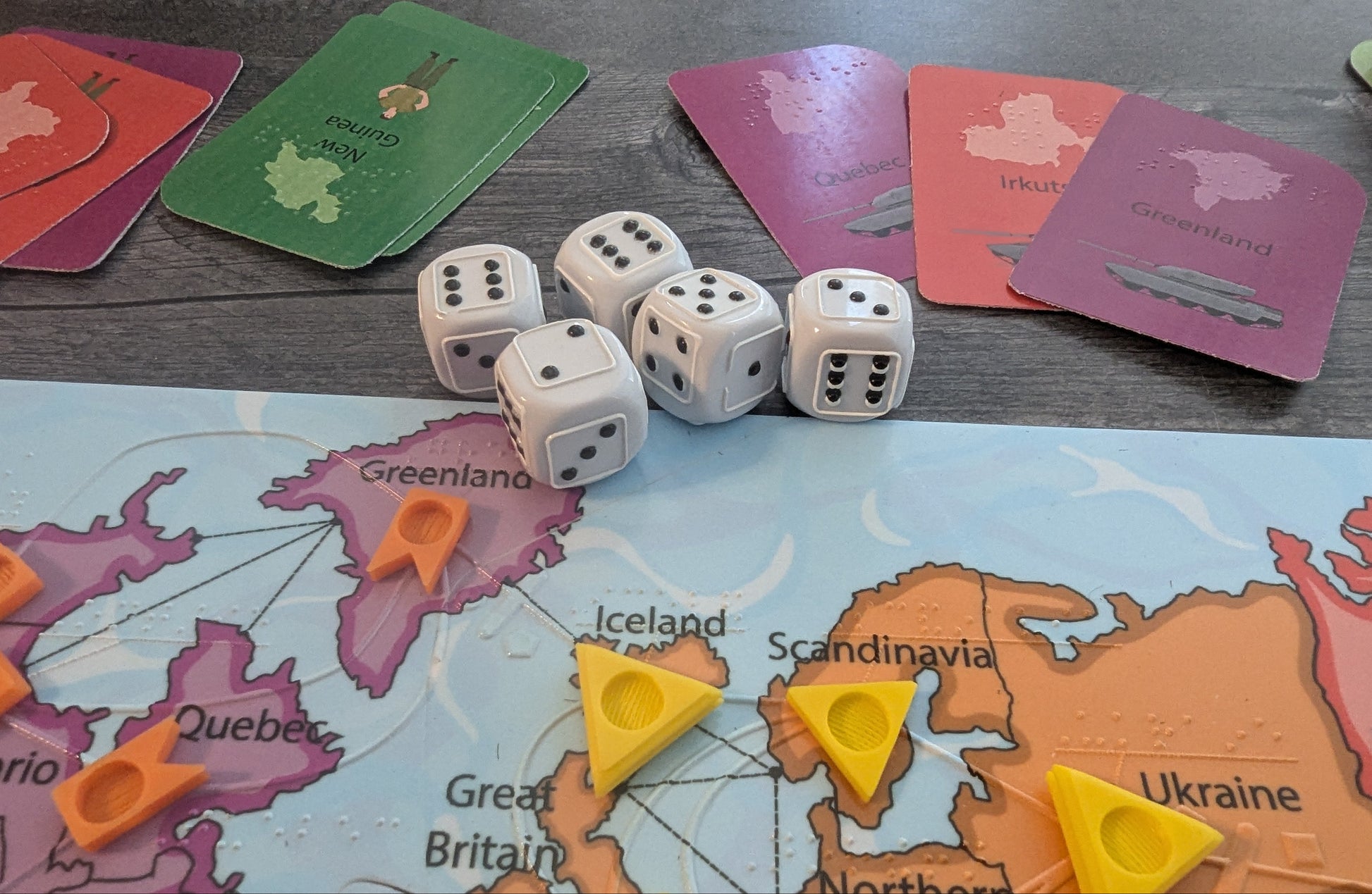 Board game setup with dice, cards, and a map of Europe on a wooden surface. The cards have braille on them and the dice are tactile.
