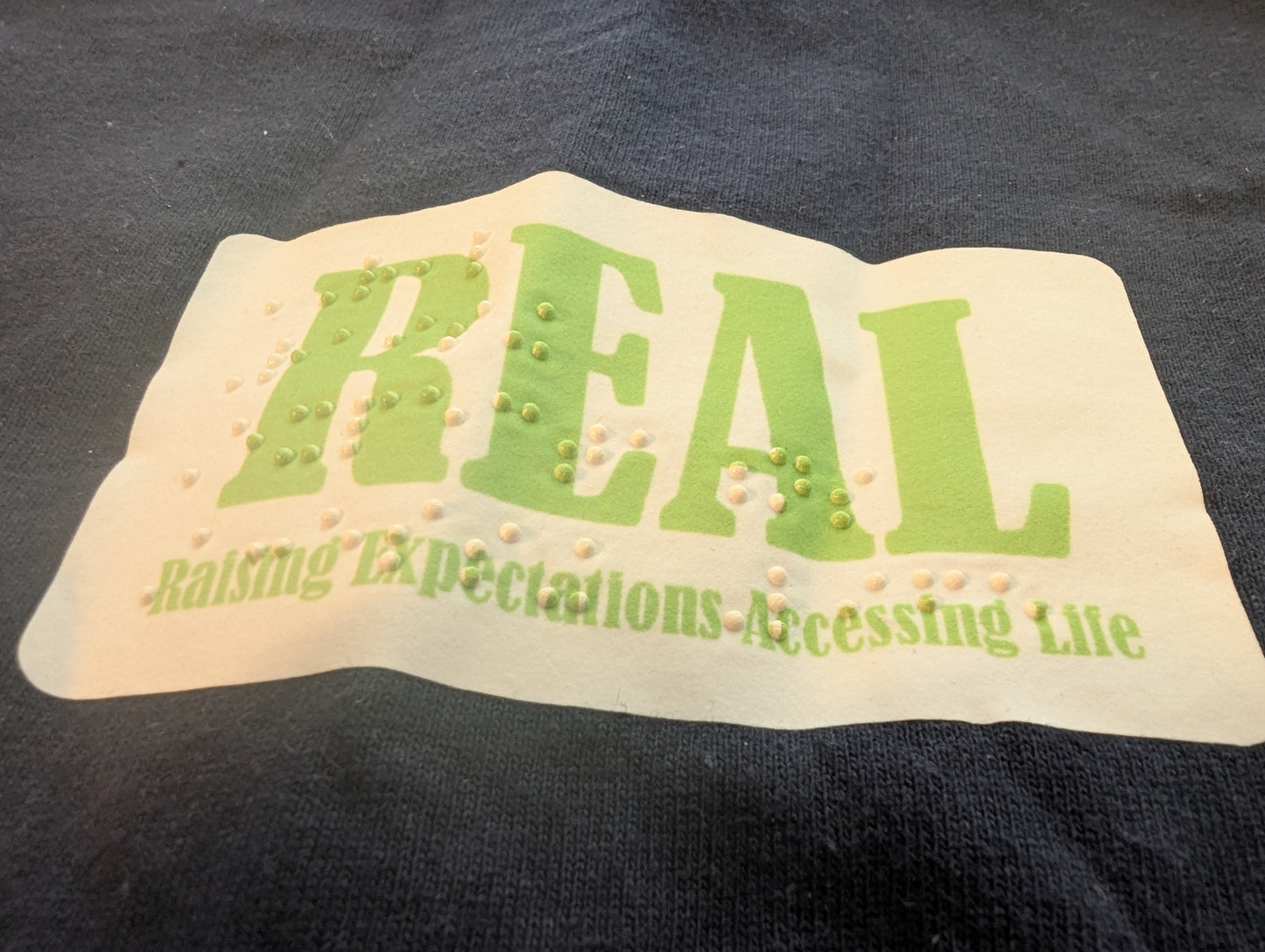 close up of braille and print text with 'REAL' text on a t-shirt