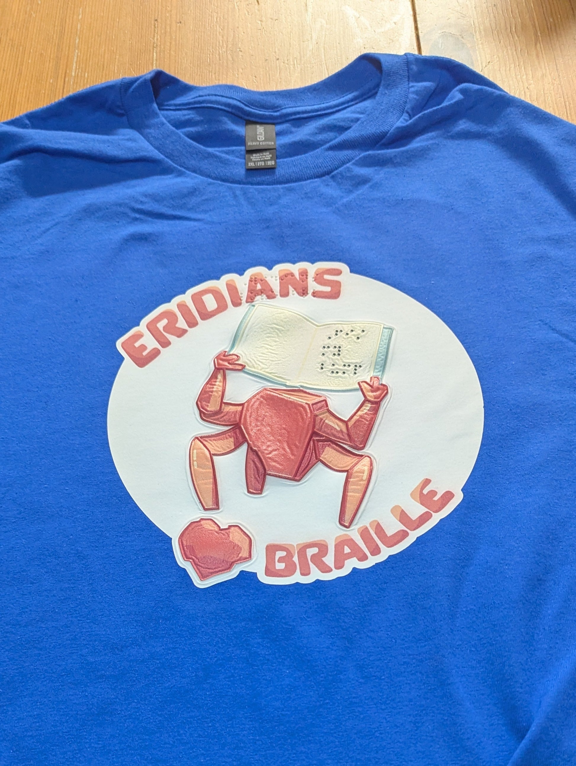 A blue t-shirt showing Rocky reading braille. The image is tactile and can be felt. It also includes braille of the test "Eridians love braille" and "fist my bump" that is written on Rocky's book