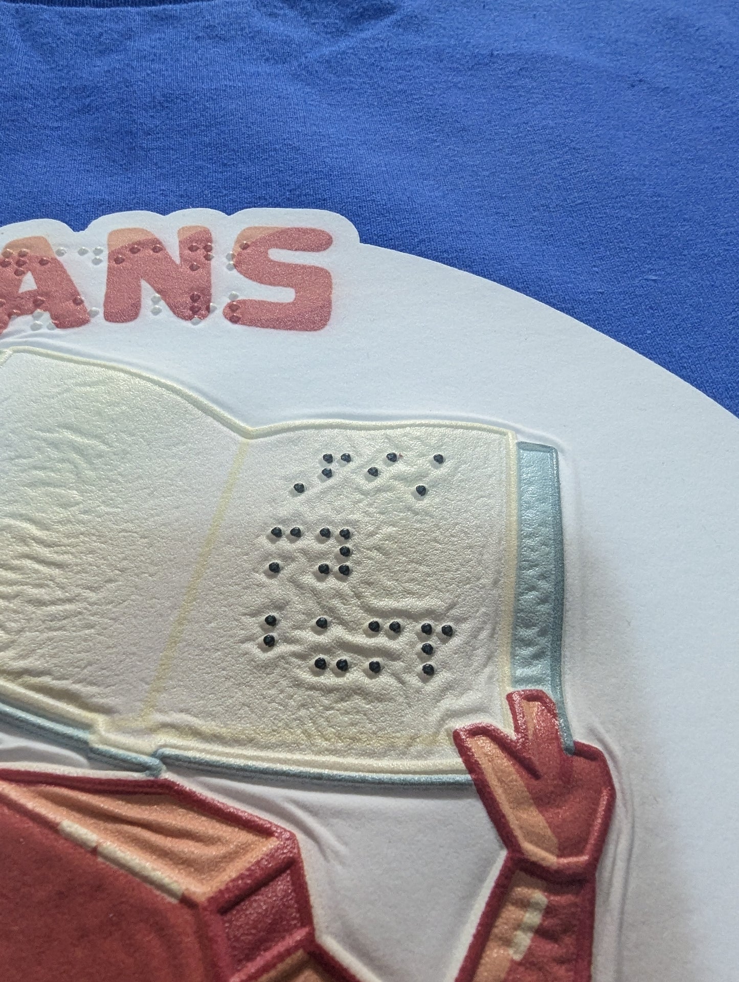 Close-up of a blue T-shirt featuring a large white graphic of a book being held by Rocky. The braille text can be seen(and felt) and reads "Fist my bump"