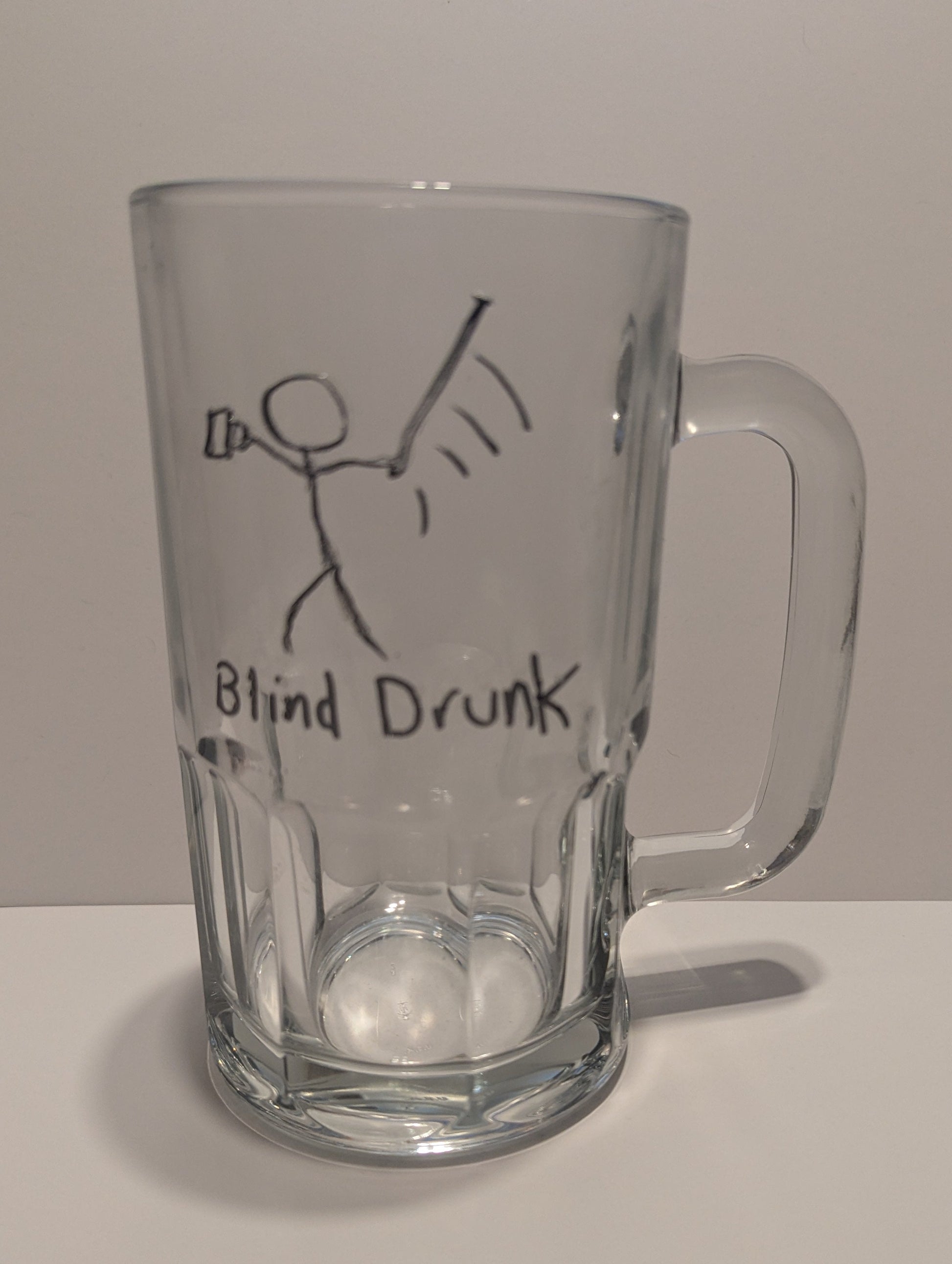 A clear glass beer mug with a handle, featuring a hand-drawn tactile black stick figure holding a mug and swinging a cane wildly. The text 'Blind Drunk' is below set against a plain light background in both print and braille.