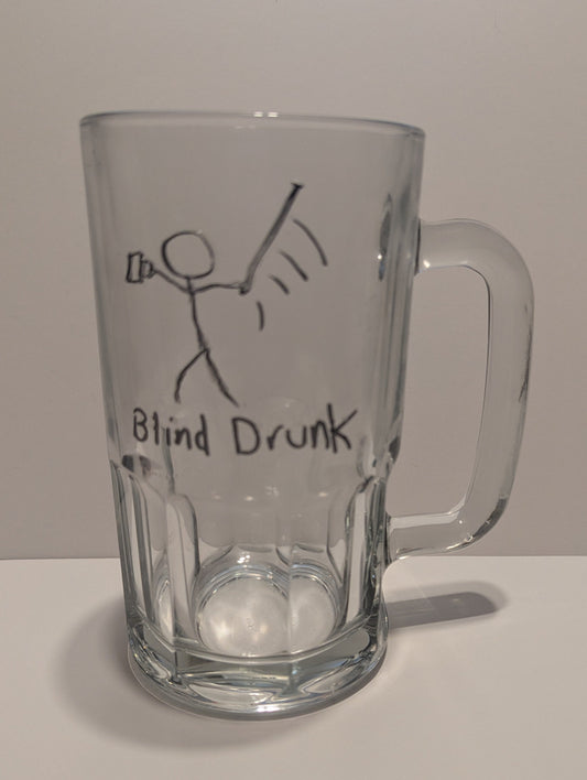 A clear glass beer mug with a handle, featuring a hand-drawn tactile black stick figure holding a mug and swinging a cane wildly. The text 'Blind Drunk' is below set against a plain light background in both print and braille.