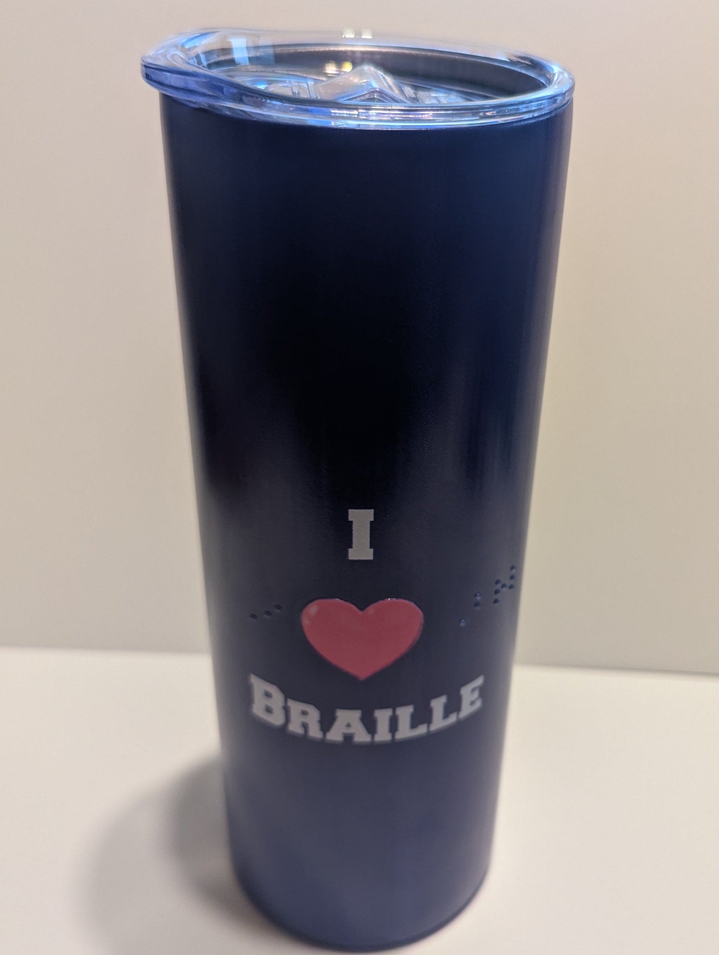 A navy blue insulated tumbler with a clear lid, featuring a graphic that reads 'I' above a pink heart and the word 'BRAILLE' in white, set against a plain light background.