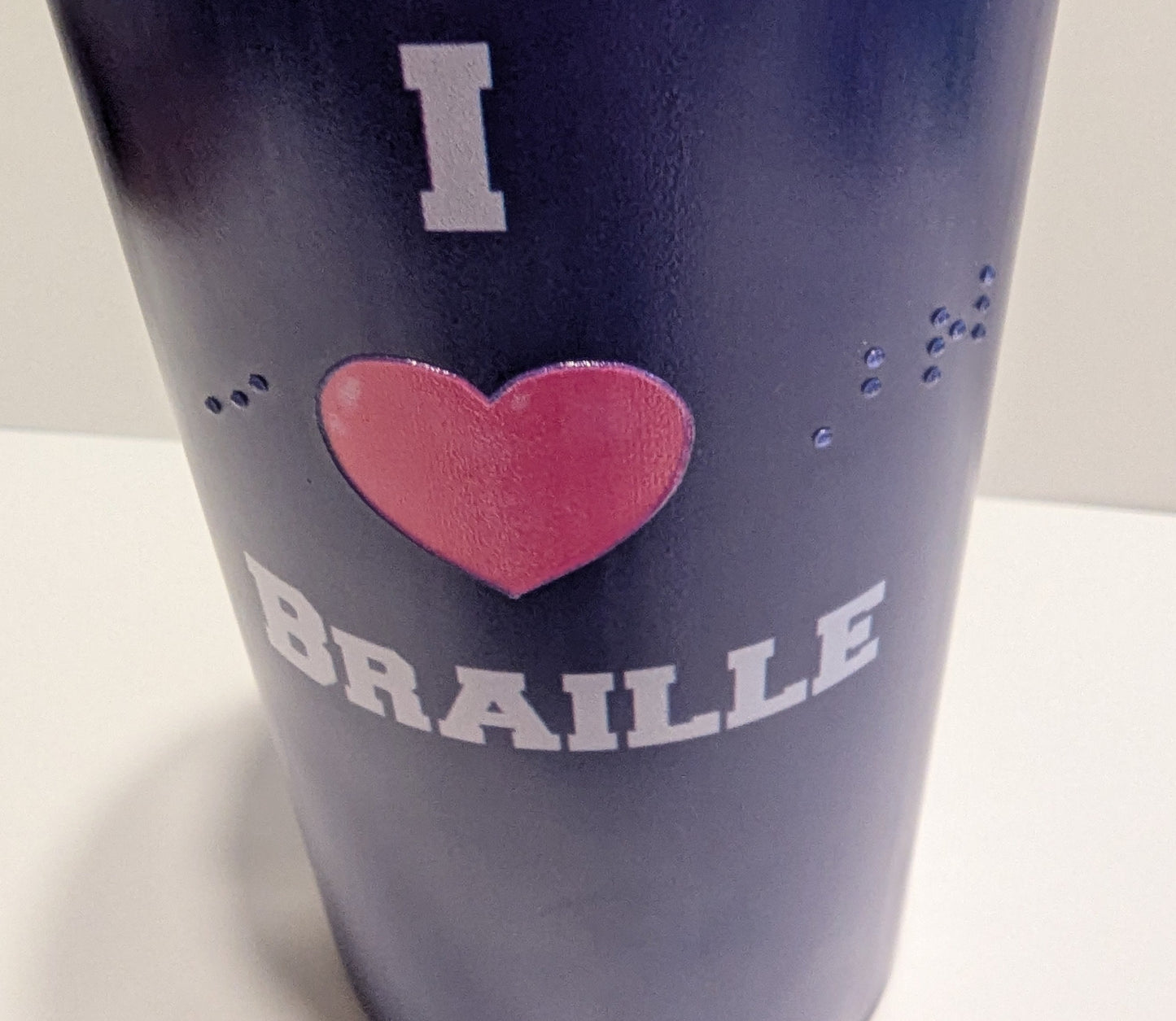 A close-up of a dark blue insulated tumbler with a pink heart and white text reading 'I BRAILLE' on a plain light background.