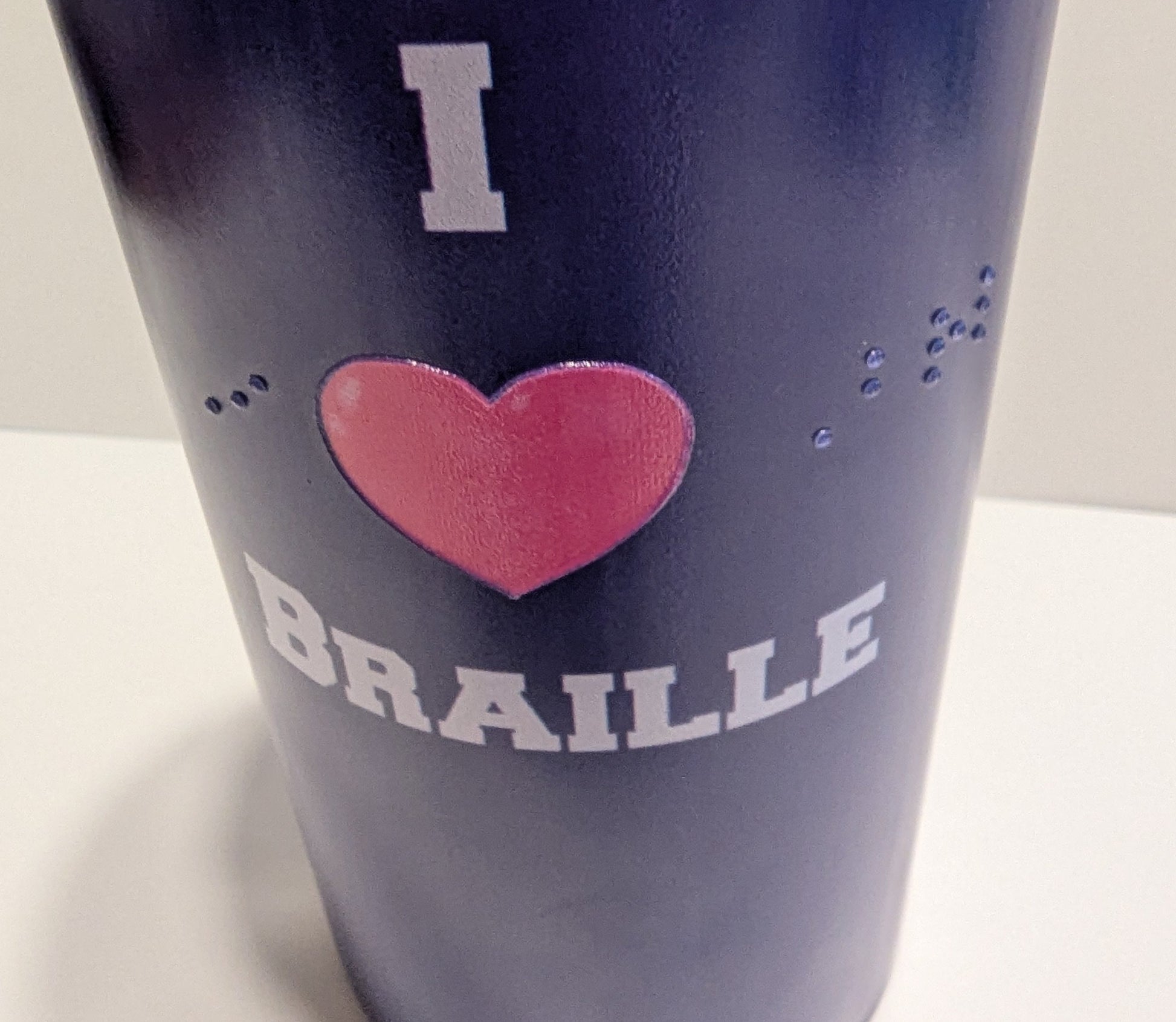 A close-up of a dark blue insulated tumbler with a pink heart and white text reading 'I BRAILLE' on a plain light background.
