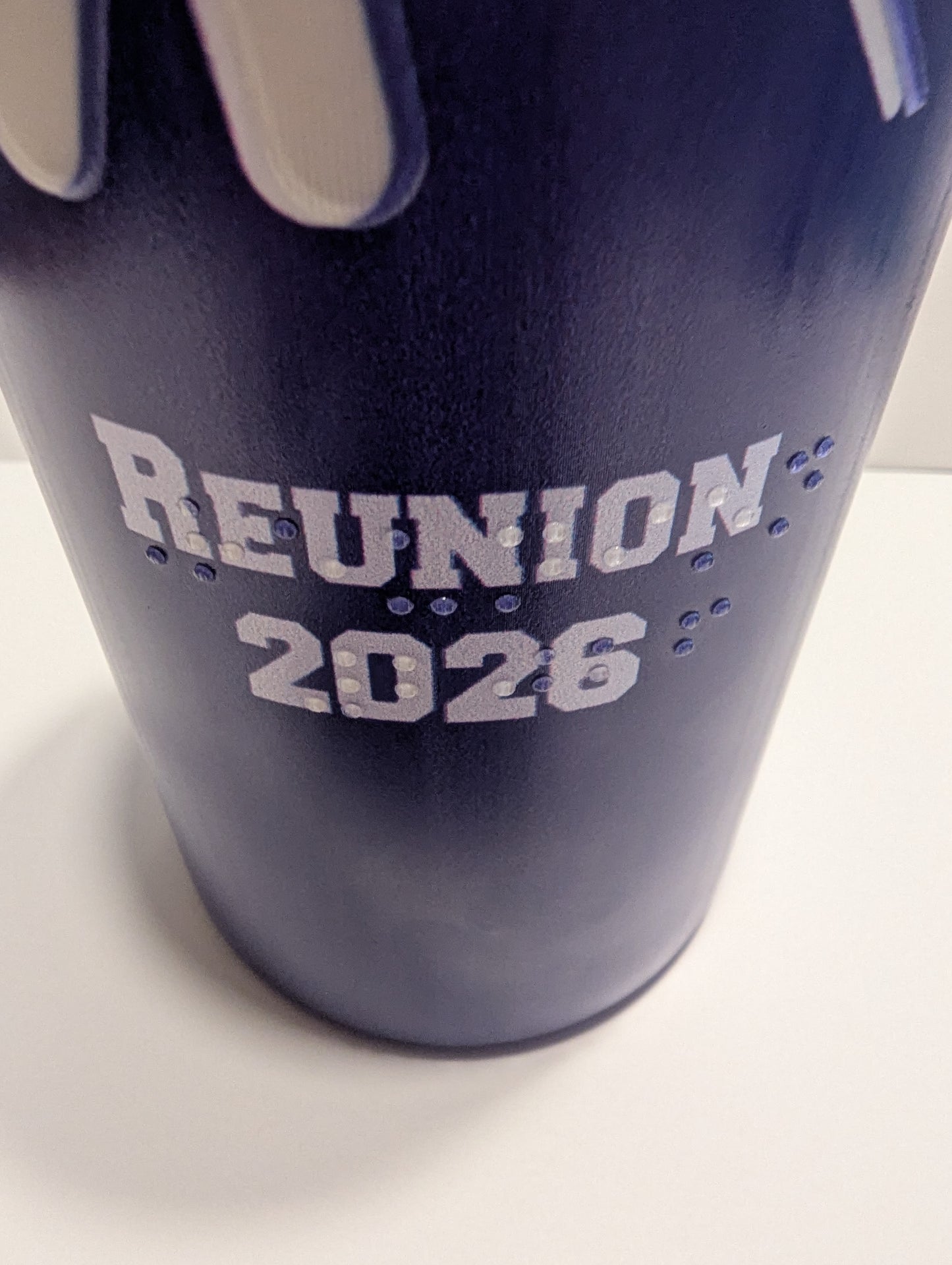 A close-up of a dark blue tumbler with white lettering that reads 'REUNION 2026' and white braille text below, photographed on a white surface.