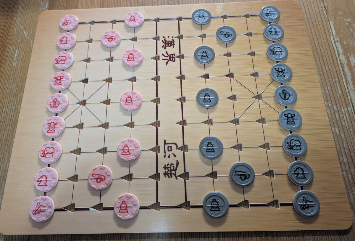 An acrylic Chinese chess board with a wooden print on a wooden table, featuring a light tan board with embossed grid lines, red and gray pieces with braille, and Chinese characters printed on the board.