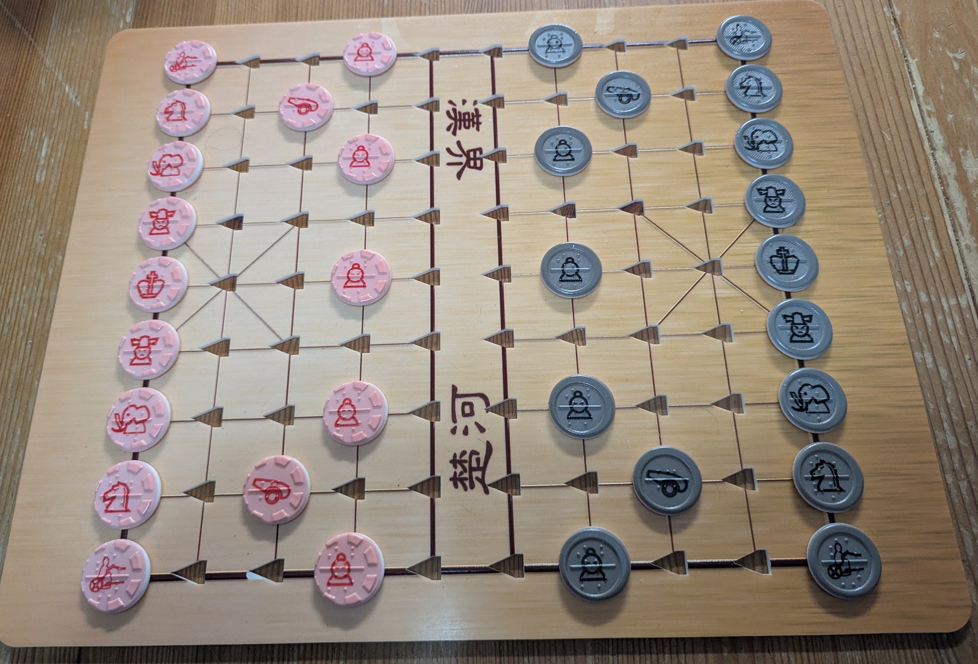 An acrylic Chinese chess board with a wooden print on a wooden table, featuring a light tan board with embossed grid lines, red and gray pieces with braille, and Chinese characters printed on the board.