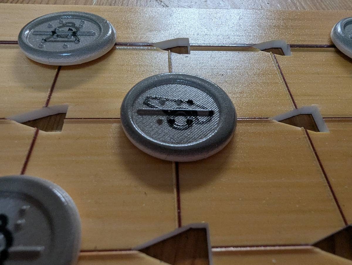 Close-up of round gray tokens placed on the squares. The central token has a cannon symbol printed on it with the letters ca in braille on the piece. There is a full border on the outside of the piece as opposed to the dotted border on the red pieces. Other similar tokens are partially visible around the board.
