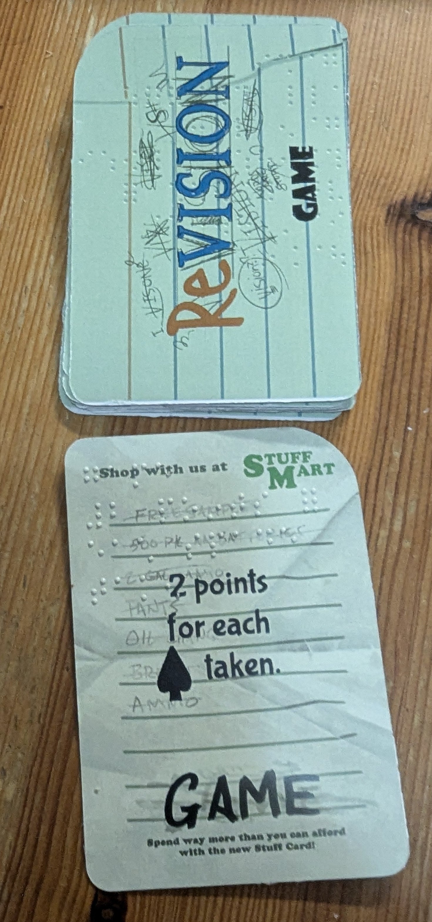 A close-up of the 'game' cards of revision on a wooden surface. One card is the back, featuring a light green background with blue lined notebook-style stripes and the word 'Revision' in red and blue. The card describes the way that players gain points this round.