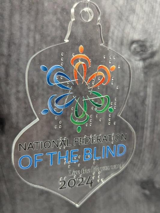 A transparent acrylic ornament with the NFB logo in print on the front. The logo is tactile. The clear braille on the back of the ornament can be seen through it. 